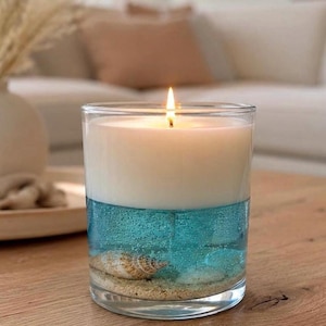Può includere: Un portacandele in vetro trasparente contenente una candela a strati con una parte superiore in cera bianca e una inferiore in gel blu traslucido. La base presenta uno strato di sabbia chiara e una conchiglia immersa nel gel blu acceso.