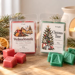 Könnte beinhalten: Zwei durchsichtige Kunststoffverpackungen mit D.Light Wachsmelts. Eine Packung mit der Aufschrift 'Weihnachtsmarkt' enthält rote Wachswürfel, die andere mit 'Christmas Tree' grüne Wachswürfel mit Tannenbaummotiv. Jede Packung wiegt 80g.