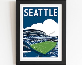 May include: A framed art print featuring a stylized illustration of a Seattle stadium with a green field, blue seating, and white clouds against a dark blue sky. The word SEATTLE is printed in large white capital letters at the top of the print.