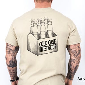 May include: A sand-colored t-shirt featuring a black ink-style graphic on the back. The illustration depicts a six-pack of glass bottles inside a cardboard carrier. The text COLD CASE INVESTIGATOR is printed in bold, black capital letters on the front.