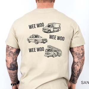 Peut inclure: Un t-shirt couleur sable avec une impression graphique noire au dos. Le motif comprend des illustrations d'une ambulance, d'une voiture de police et d'un camion de pompiers, accompagnées du texte WEE WOO en police noire et grasse.