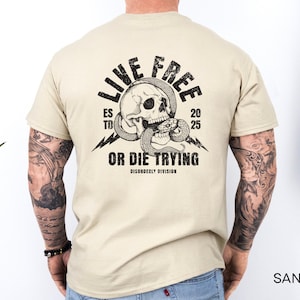 May include: Sand-colored t-shirt featuring a skull entwined by a snake, with the text "LIVE FREE OR DIE TRYING" and "ESTD 2025" in a distressed font. The design includes lightning bolts. The shirt is a casual wear item.