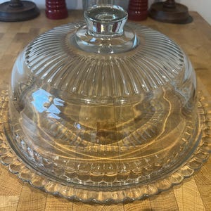 Op de afbeelding: Een heldere glazen taartstandaard met een gewelfde deksel. Het deksel heeft verticale ribbels en een kleine handgreep. De basis is geschulpt. De standaard staat op een houten oppervlak. Een decoratief stuk voor het serveren van taarten en gebak.