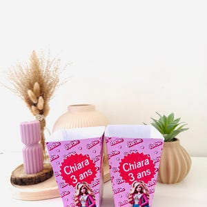 Peut inclure: Deux boîtes à cadeaux roses sur le thème de Barbie. Chaque boîte présente le nom "Chiara" et l'âge "3 ans" dans un motif d'éclat d'étoile rouge. Les boîtes sont décorées avec des logos Barbie et des images de Barbie.