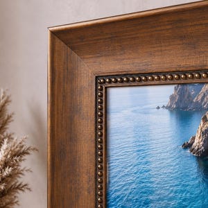 May include: A rectangular picture frame with a dark brown, wood-grain finish and a decorative inner border featuring a row of small, raised metallic beads. The frame encloses a photograph of a rocky cliffside overlooking deep blue ocean water.