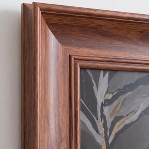 Puede incluir: Un marco rectangular con un acabado de grano de madera marrón medio pulido y bordes biselados. El marco encierra una impresión gris oscuro con formas abstractas y fluidas similares a hojas en tonos blancos, crema y acentos dorados.