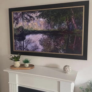 May include: A rectangular landscape painting in a black frame with a gold inner border, depicting a dark pond reflecting trees and foliage in shades of green, purple, and brown. Below the painting are two small potted plants and a patterned sphere.