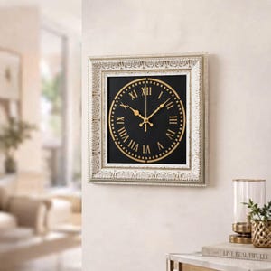 May include: A square wall clock featuring an ornate, distressed white frame with intricate carvings. The clock face is black with gold-colored Roman numerals and matching gold hands, set within a circular gold border on a square background.