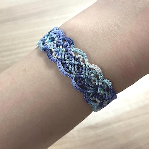 May include: A woven macrame bracelet featuring an intricate knotted pattern in a gradient of blue, navy, and light turquoise threads. The design consists of repeating scalloped edges and interconnected loops that form a continuous band.