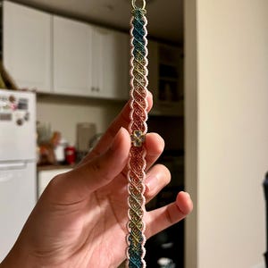 May include: A macrame bracelet in a gradient of colors, including blue, green, yellow, orange, and red. The bracelet features a woven design with a small, decorative charm. The bracelet is held up against a blurred background.