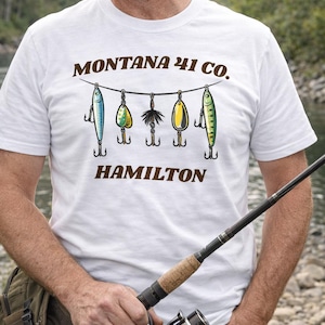May include: White t-shirt with the text "MONTANA 41 CO. HAMILTON" in brown. The shirt features a graphic of fishing lures hanging from a line. A fishing rod is held in the person's hand.