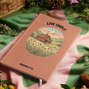 Puede incluir: Un cuaderno color melocotón con las palabras "LIVE SIMPLY" y "MONTANA 41 Co." impresas en la portada. La portada presenta una ilustración circular de un granero rojo en un campo. Una cinta blanca como marcapáginas es visible.