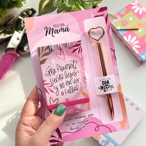 Op de afbeelding: Een roze cadeau set voor Moederdag, met een notitieblok met de tekst "Me Prometi LLegar Lejos y No me voy a Fallar", een pen met hartvormige top en de woorden "Feliz Día Mamá". De set wordt in een hand gehouden.
