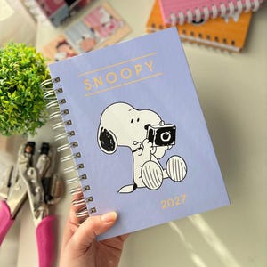 Pode incluir: Uma agenda de espiral em tom lilás claro com um gráfico do Snoopy segurando uma câmera. O texto "SNOOPY" está impresso em amarelo no topo e o ano "2027" está impresso em amarelo no canto inferior direito da capa frontal.