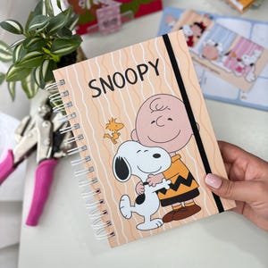 Puede incluir: Un cuaderno de espiral con una cubierta de color melocotón claro que muestra a Snoopy abrazando a Charlie Brown y Woodstock. El texto SNOOPY está impreso en negro en la parte superior. Una banda elástica negra vertical asegura la cubierta con ondas.