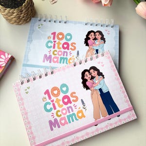 以下が含まれることがあります： 「100 Citas con Mama」の文字と、抱き合う女性2人のイラストが描かれた、スパイラル綴じのノート2冊。ノートはピンクとブルーの表紙で、装飾的なボーダーとゴムバンド留めが施されています。背景には、編み込みの鉢植えとピンクのチューリップがあります。