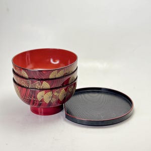 May include: A stack of three red and black lacquered bowls with a matching black tray. The bowls feature a gold fan design and a glossy finish. The tray has a black wood grain pattern.