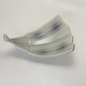 Puede incluir: Un plato de cerámica en forma de hoja, de color gris claro, con una textura ondulada. El plato tiene un sutil degradado de tonos morados y azules. El borde está delineado con un acabado metálico plateado. El plato está diseñado con una forma curva y asimétrica.