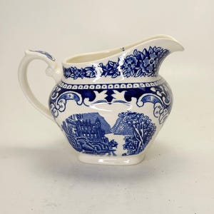 May include: A white ceramic pitcher with a blue and white landscape design. The pitcher features a handle and a spout. The design includes buildings, trees, and floral patterns. The pitcher is likely for serving liquids.
