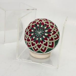 Puede incluir: Una bola Temari artesanal con un diseño geométrico complejo en tonos verdes, rojos y blancos. La bola se exhibe sobre un pequeño soporte de madera dentro de una caja de acrílico transparente, mostrando el intrincado bordado.