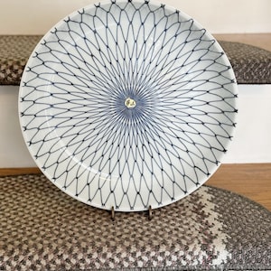 May include: A white ceramic plate with a blue geometric pattern. The plate is round and has a delicate, web-like design radiating from the center. It is displayed on a metal stand, showcasing its intricate details. The plate is a decorative item.