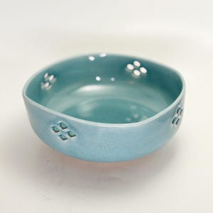 May include: A light blue ceramic bowl with a crackle glaze finish. The bowl has a rounded shape with decorative cut-out patterns. The cut-outs are arranged in a diamond and dot pattern around the rim of the bowl. The bowl is empty and sits on a white surface.