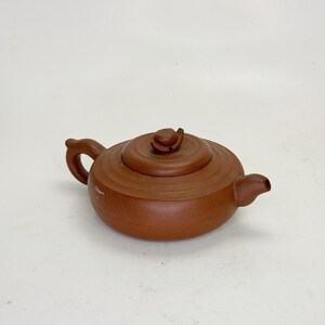 May include: A small, brown ceramic teapot with a rounded body, a curved handle, and a spout. The lid has a small knob. The teapot is set against a plain white background. The teapot appears to be made of clay.