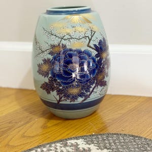 May include: A light blue ceramic vase with a dark blue rim and base. The vase features a large, detailed blue peony flower design with gold accents. The top of the vase has a gold mountain and tree design.