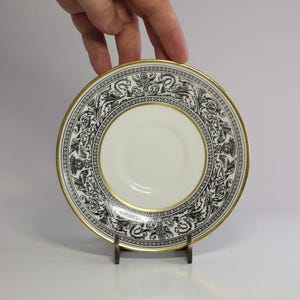 May include: A white saucer with a black and gold decorative border. The saucer has a gold rim and a central well. The border features a repeating pattern of stylized figures and floral elements. The saucer is held by a hand.