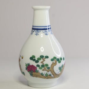 May include: White ceramic vase with a narrow neck and a rounded body. The vase has a blue band with a decorative pattern near the top. The body is decorated with a painted scene of a tree, flowers, and a butterfly.