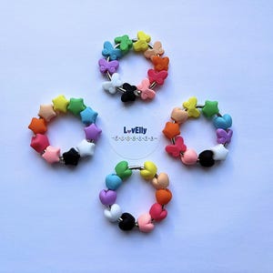 May include: Four colourful beaded rings with various shapes, including butterflies, stars, and hearts. Each ring features a different colour scheme, with a mix of rainbow hues and black and white accents. The rings are arranged on a white surface, with the text "LovElly" visible.