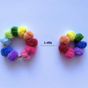 May include: Two rainbow pom-pom bracelets. Each bracelet is a circle of fluffy pom-poms in a spectrum of colours, including red, pink, orange, yellow, green, blue, and purple. The bracelets are on a white background.