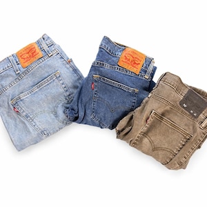 Peut inclure: Trois paires de jeans en denim pliés, bleu clair, bleu foncé et marron. Chaque paire présente un patch en cuir rouge et beige sur la poche arrière. Les jeans sont disposés sur fond blanc.