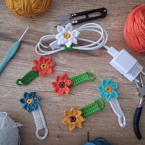 Peut inclure: Plusieurs organiseurs de câbles en forme de fleurs crochetées à la main, de couleurs variées : blanc, orange, jaune et bleu. L'image présente également de la laine, des ciseaux et un crochet, suggérant un thème artisanal.