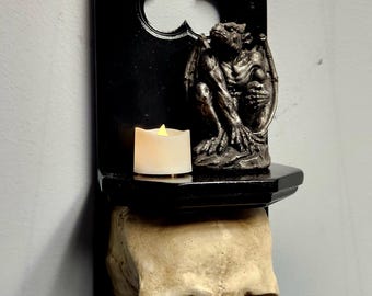 May include: A black wall-mounted shelf with a pointed gothic arch backplate. A small grey gargoyle figurine and a white LED tea light candle sit on the shelf. The base of the shelf is supported by a realistic human skull sculpture in off-white.