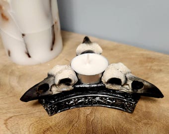 May include: A decorative candle holder featuring a central tea light surrounded by three raven skull replicas. The skulls are off-white with black beaks, set on a black, ornate base. A large white candle is in the background.