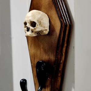 May include: A wall-mounted hook featuring a dark brown, stained wooden backboard shaped like a coffin. An off-white, realistic human skull replica is attached to the upper portion of the wood, positioned above a curved, matte black metal hook.