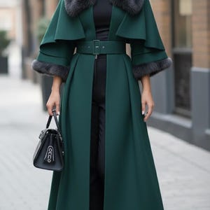 May include: A long, emerald green coat with a matching belt and fur trim. The coat features a cape-like design and is paired with black pants, a black hat, and a black handbag. The outfit is completed with black boots.
