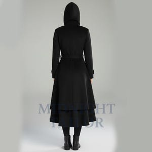 以下が含まれることがあります： フード、ベルト、フレアスカートが付いた黒のロングコート。コートは二層構造のスカートデザインで、黒のレギンスとブーツを着用しています。背面図は、コートのテーラードシルエットと詳細を示しています。