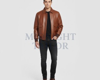 May include: A brown leather bomber jacket with a collared neckline and two front pockets. The jacket is worn with a black t-shirt and dark wash jeans. The model is wearing brown leather boots.