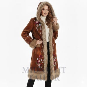 May include: A long brown coat with a faux fur-lined hood and trim. The coat features embroidered floral designs in pink, blue, and green. The coat has long sleeves and a button-down front. The model is wearing a white turtleneck underneath.