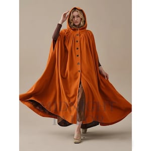 Op de afbeelding: Een oranje cape met capuchon en een knoopsluiting aan de voorkant. De cape is gemaakt van een zachte stof en heeft een vloeiend ontwerp. Het model draagt er een bruin shirt met lange mouwen onder. De cape is een herfstmode-item.