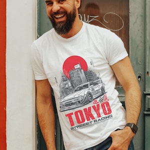Puede incluir: Camiseta blanca con un diseño gráfico de un coche, un paisaje urbano y un sol rojo. La palabra "TOKYO" está en rojo y "STREET RACING" en blanco. El diseño está en la parte delantera de la camiseta.