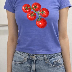 May include: A blue t-shirt featuring a watercolor illustration of five ripe red tomatoes on a green vine. The tomatoes are clustered together, with visible details of their shape and texture. The shirt is paired with blue denim jeans.