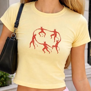 May include: Yellow short-sleeved t-shirt with a red graphic of six figures holding hands in a circle. The shirt is a light yellow color and has a crew neck. The graphic is a simple, stylized design.