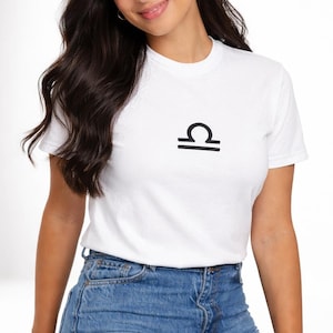 Könnte beinhalten: Weißes T-Shirt mit einem schwarzen Waage-Sternzeichen. Das kurzärmelige Oberteil ist aus einem weichen Material gefertigt und wird mit blauen Jeans kombiniert. Das Waage-Symbol ist mittig auf der Brust platziert.