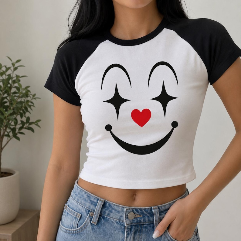 May include: White crop top with black sleeves and a black collar. The shirt features a large clown face design with black star-shaped eyes, a red heart nose, and a smiling mouth. The shirt is made of a soft, comfortable material.