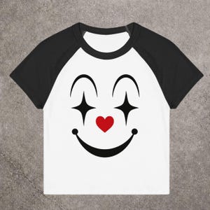 May include: White and black raglan t-shirt with a clown face design. The design features black arched eyebrows, diamond-shaped eyes, a red heart nose, and a smiling mouth. The shirt has black sleeves and a white body.