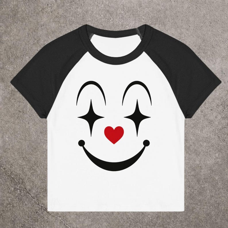 May include: White and black raglan t-shirt with a clown face design. The design features black arched eyebrows, diamond-shaped eyes, a red heart nose, and a smiling mouth. The shirt has black sleeves and a white body.