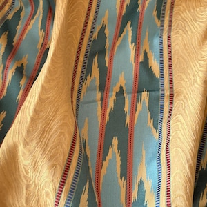 May include: Close-up of a textured fabric with a golden-yellow background. Vertical stripes in teal, light blue, red, and gold create a patterned design. The fabric has a subtle, wavy texture.